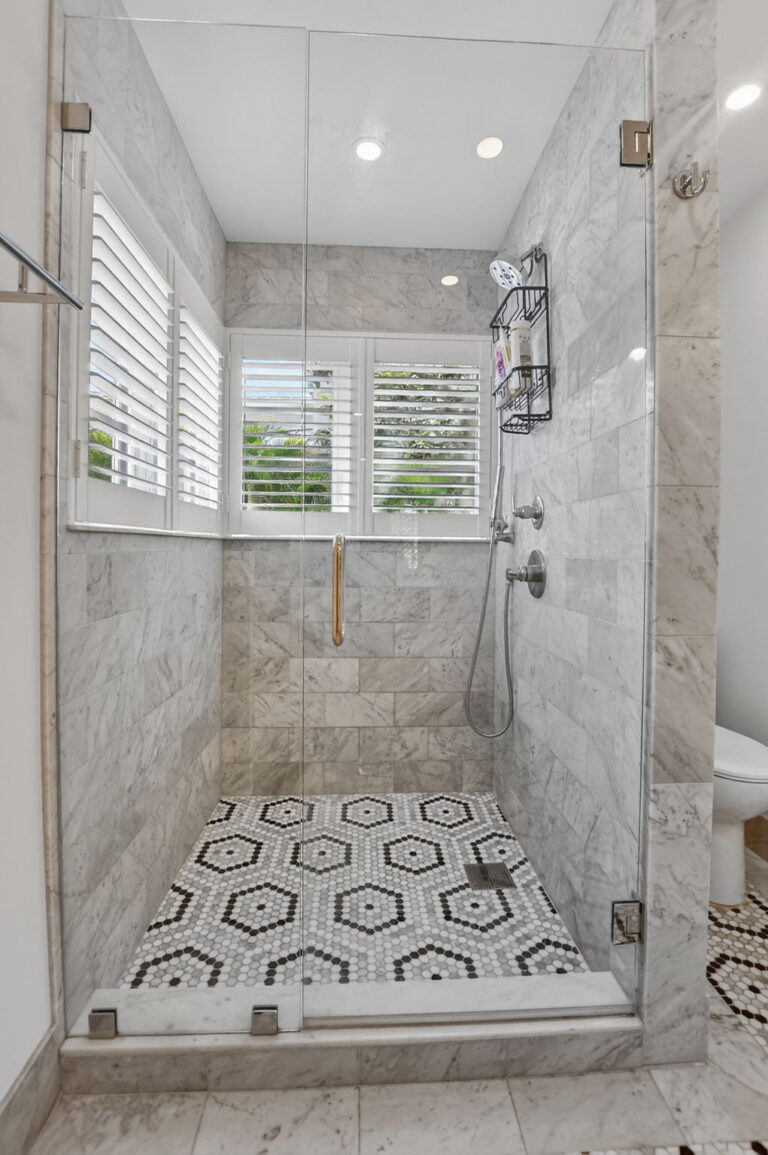 Walk-in shower with marble tile walls, glass enclosure, chrome fixtures, hexagonal floor pattern, and plantation-shutter window.