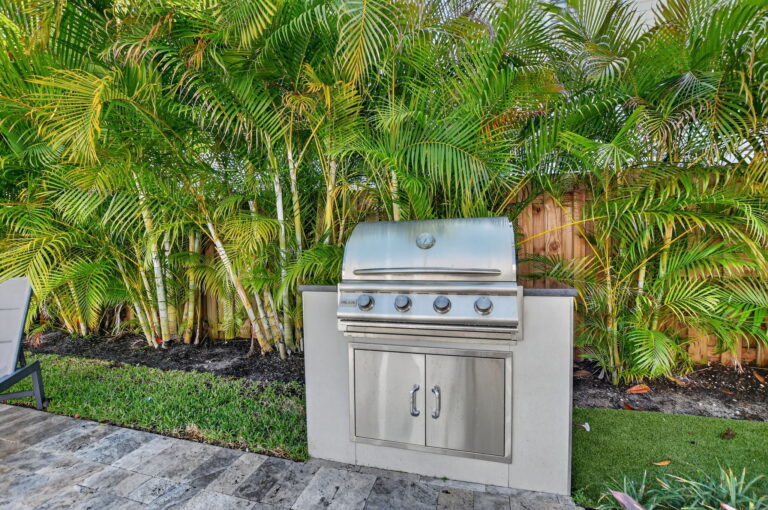 Outdoor kitchen area with a built-in stainless steel gas grill on a stone counter, set against palm trees and a wooden fence.