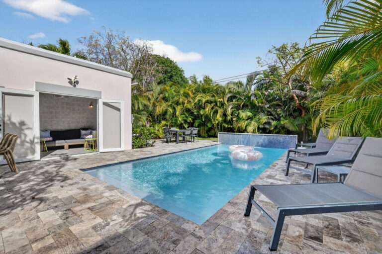 Pool area with open pool house doors revealing an indoor lounge, stone patio, lounge chairs, and lush tropical greenery.