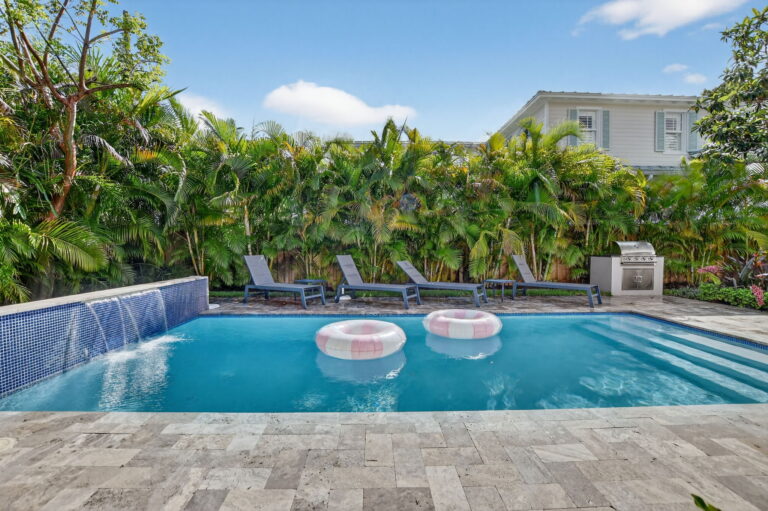 Rectangular swimming pool with blue mosaic waterfall feature, stone patio, grey lounge chairs, and tropical palm landscaping around a white rental home.