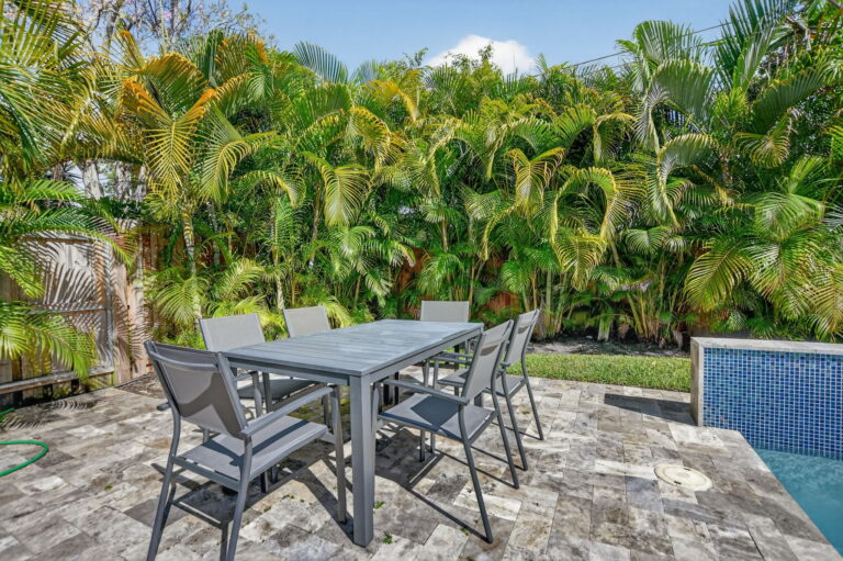 Outdoor dining area with a grey table and six chairs on a stone patio, with palm trees in the background and a swimming pool nearby.