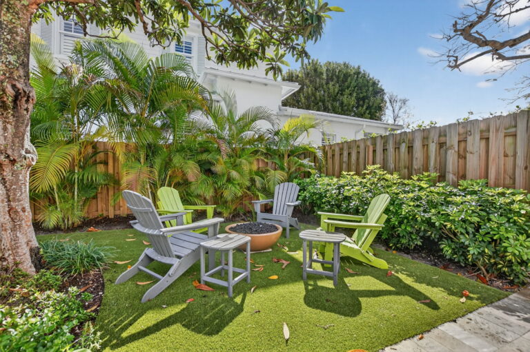 Private backyard seating area with Adirondack chairs around a fire pit on artificial grass, surrounded by a wooden fence and tall palm trees.