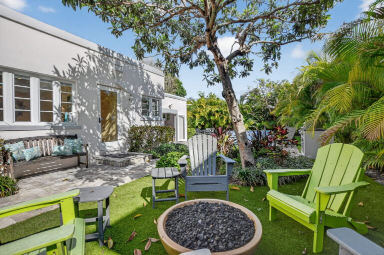 Outdoor patio and garden at a white one-story rental home, with a wooden bench, fire pit, Adirondack chairs, and tropical landscaping under a clear blue sky.