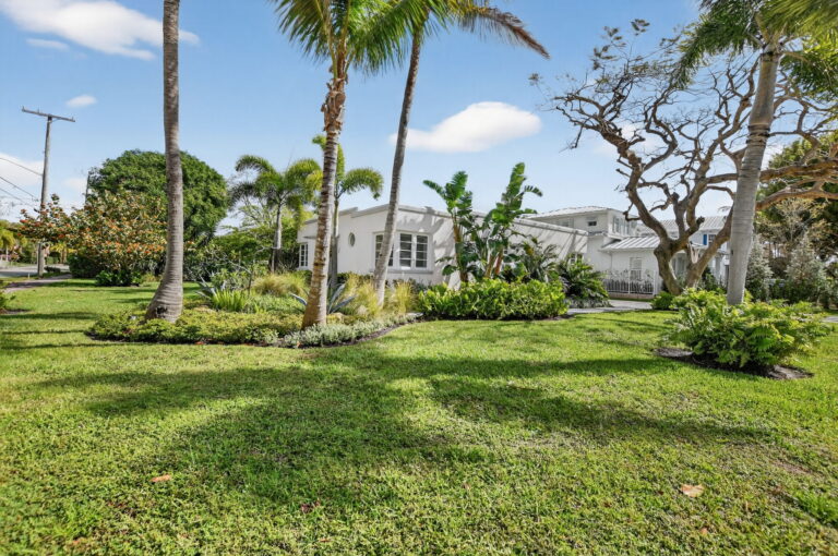 Angled exterior view of the white cottage showing the side yard, mature trees, palm landscaping, and expansive green lawn.