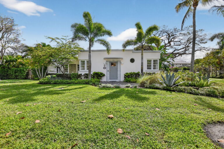 Front view of a white single-story rental cottage with grey front door, palm trees, tropical landscaping, and a wide green lawn under a blue sky.