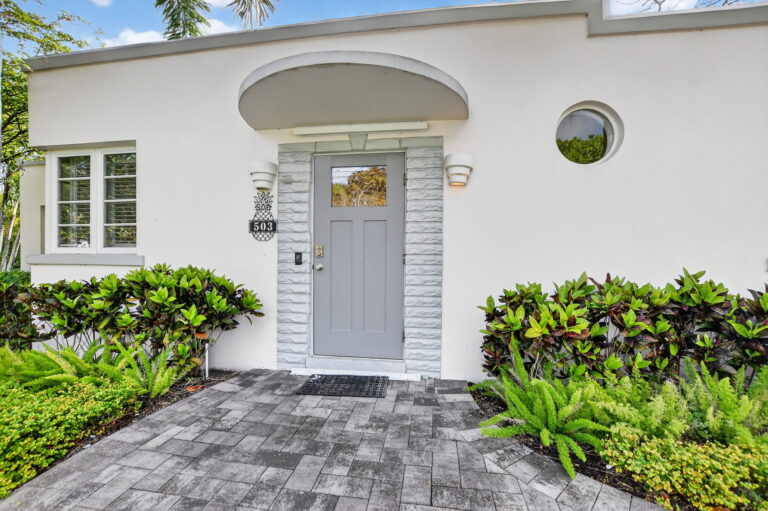 Front entrance of a modern white rental home with a grey door, stone walkway, curved overhang, and landscaped garden beds.
