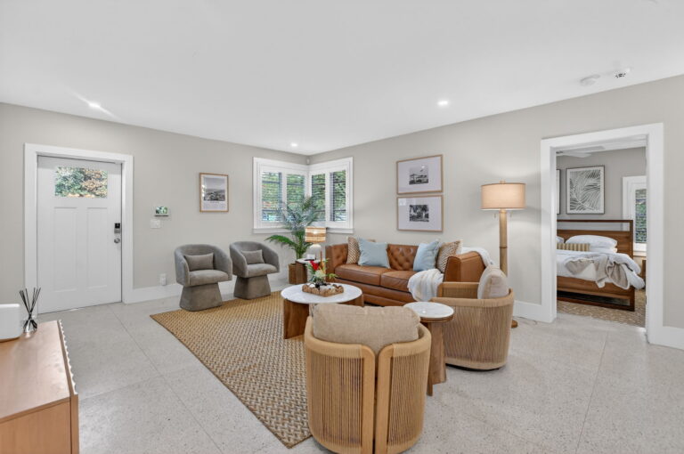 Wide view of the living room with tan leather sofa, wicker armchairs, marble coffee table, jute rug, grey accent chairs by the front door, and an open doorway to the bedroom.