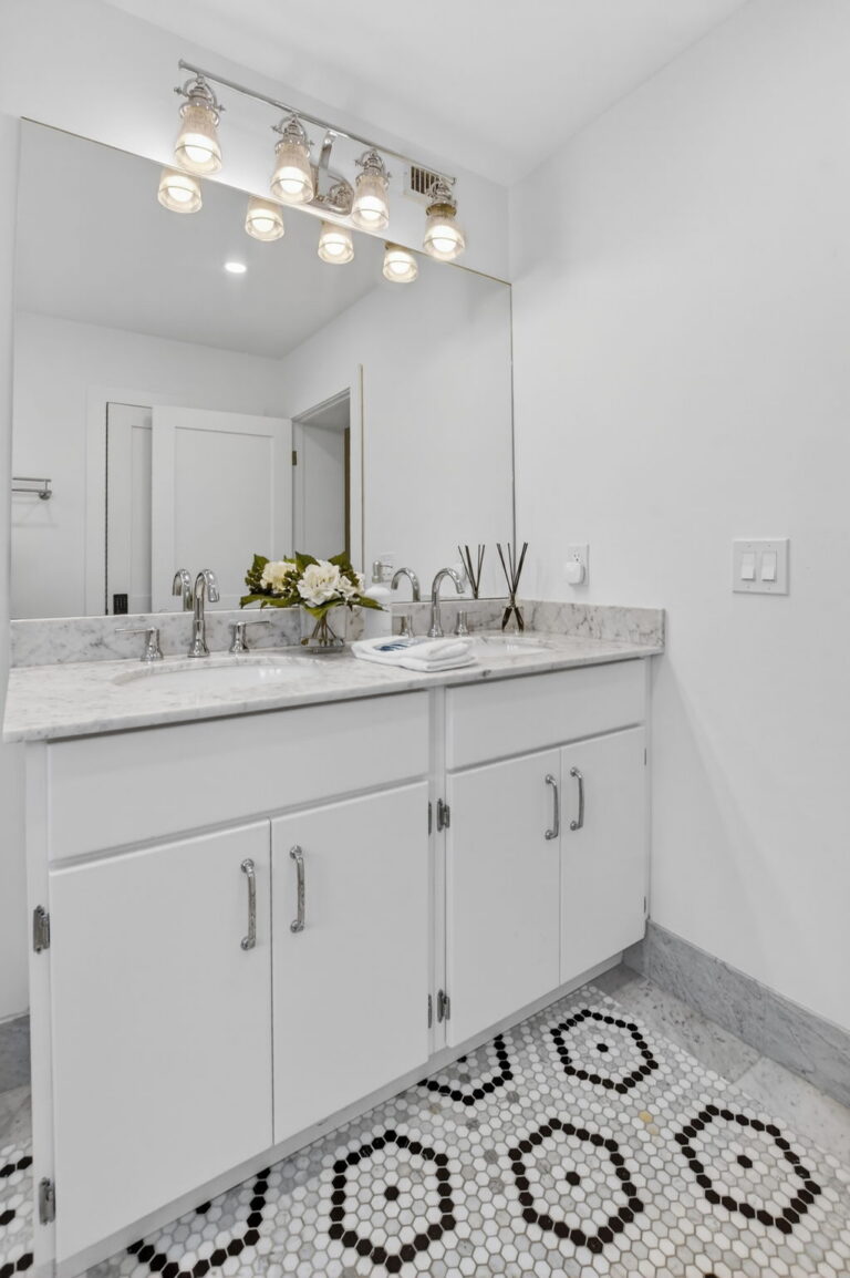 Bright bathroom with white double vanity, grey-veined marble countertop, frameless mirror, decorative light fixture, and black-and-white hexagonal floor tile.