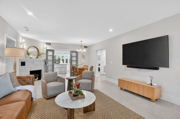 Open-concept view from the living room through the dining area to the bright white kitchen, with stone fireplace, TV wall, and French doors in the background.