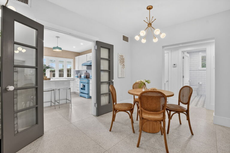 Wide view of the dining area opening to the kitchen through grey French doors, with light blue retro-style appliances, hallway, and bathroom in the background.