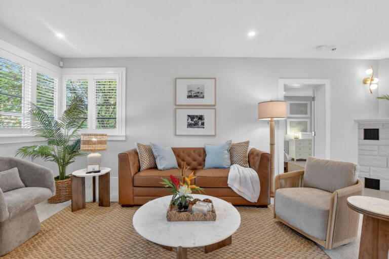 Close-up of the living room seating area with leather sofa, blue-and-white pillows, marble coffee table, potted palm plant, coastal wall art, and a glimpse of the stone fireplace.