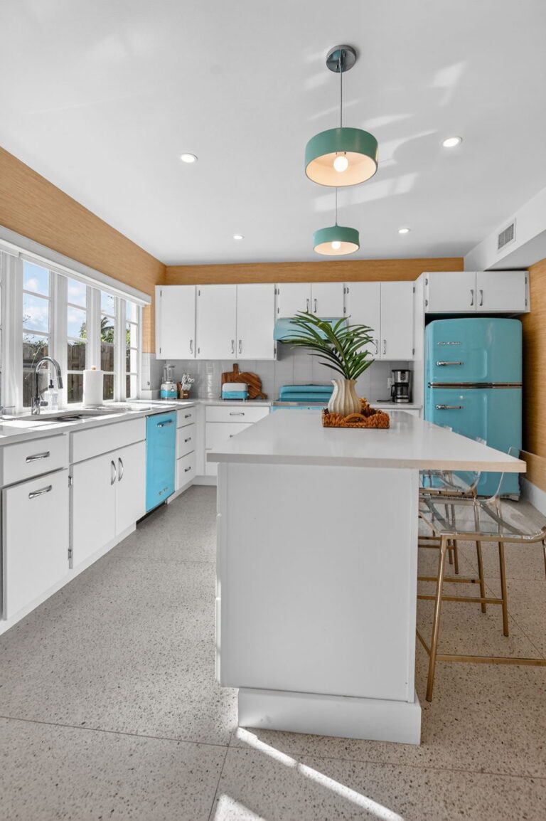 Wide view of the white kitchen with central island, clear acrylic bar stools, light blue Big Chill appliances, blue pendant lights, and garden view through the sink window.