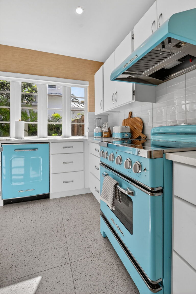 Kitchen close-up of turquoise Big Chill dishwasher and range with chrome details, white cabinets, and garden view through the window above the sink.