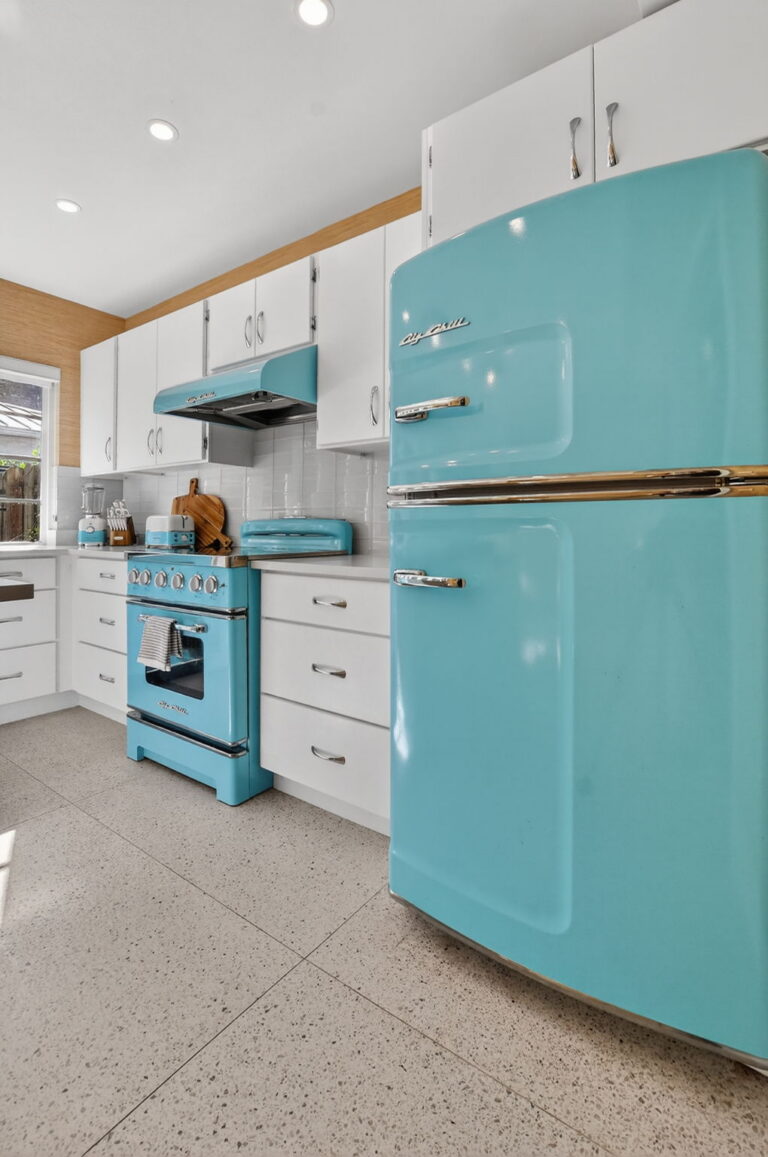 Bright white kitchen with turquoise Big Chill retro refrigerator, range, and vent hood, white cabinetry, subway tile backsplash, and terrazzo flooring.