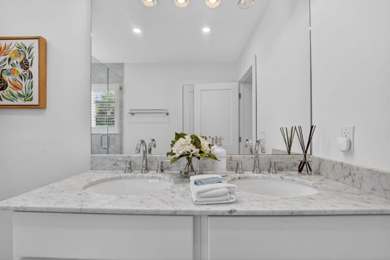 Close-up of double vanity with grey marble countertop, two sinks, chrome faucets, folded towels, floral arrangement, and colorful toucan wall art.