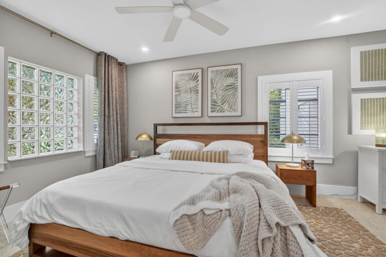 Primary bedroom with dark wood bed, white bedding, cream knit throw, palm frond art, glass-block window, and plantation shutters.