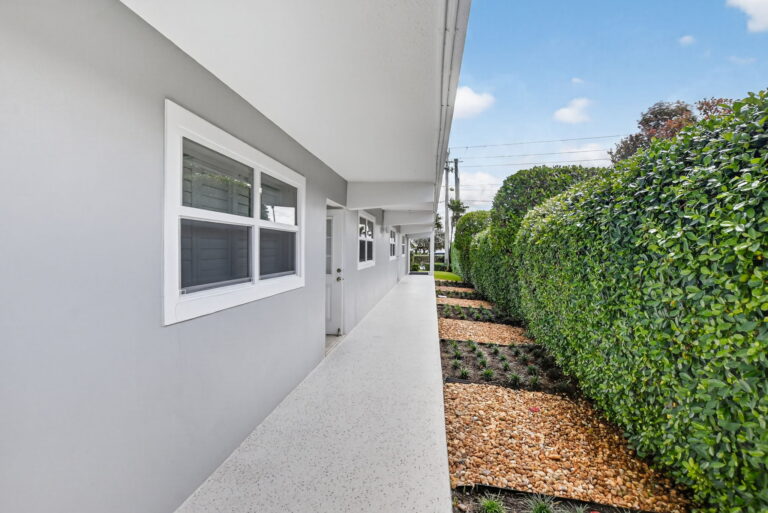 Covered exterior walkway with grey walls, white ceiling, speckled floor, privacy hedge, and landscaped garden bed.