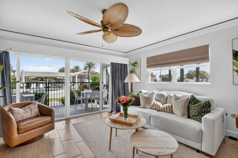 Bright living room with white sofa, wood nesting coffee tables, cognac leather swivel chair, palm-leaf ceiling fan, and sliding doors to patio.