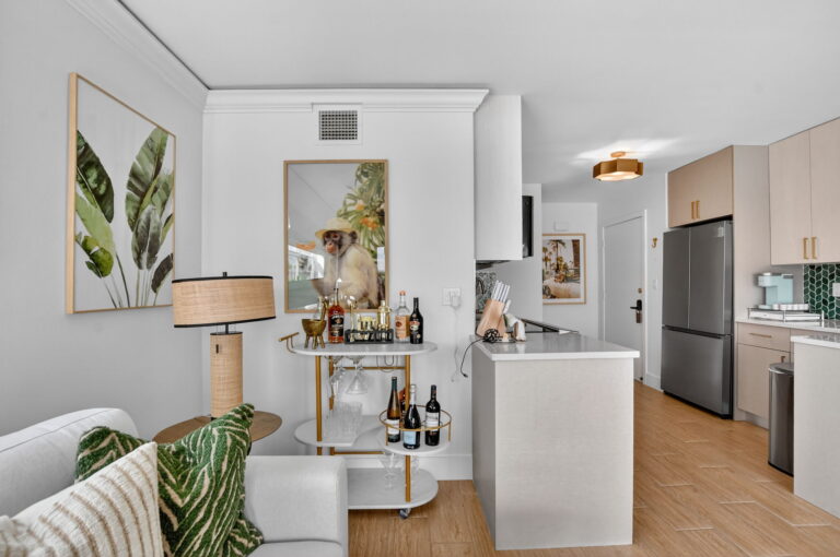 Living area flowing into kitchen with gold bar cart, tropical wall art, light wood cabinets, stainless refrigerator, and white breakfast bar.