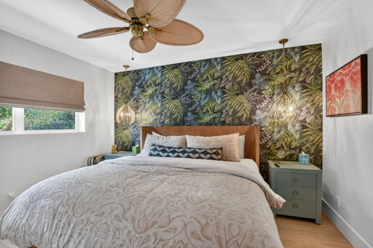 Bedroom with slat wood headboard, tropical leaf accent wallpaper, palm-leaf ceiling fan, gold pendant lights, and light blue nightstand.