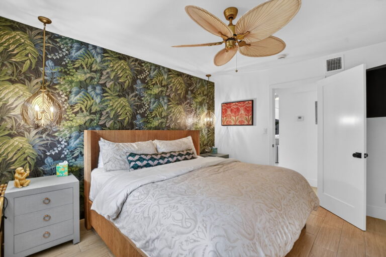 Angled bedroom view with tropical wallpaper, palm-leaf fan, gold pendant light, floral wall art, and open doorway to hallway.