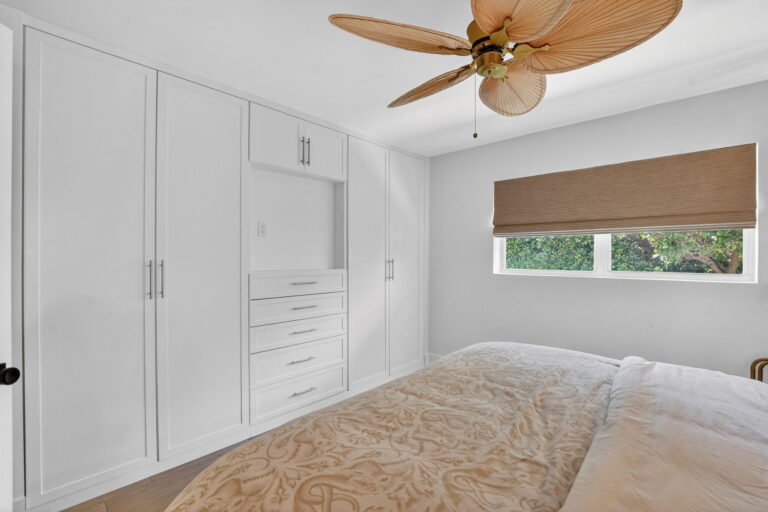 Bedroom view showing floor-to-ceiling white built-in closets, drawers, bed, woven wood blind, and palm-leaf ceiling fan.