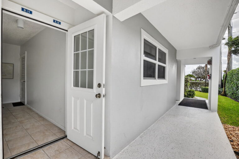 Open white front door to Delray Cabana Condo unit, tiled entry hallway, covered grey-and-white exterior walkway, and view of lawn, hedges, and palm trees beyond.