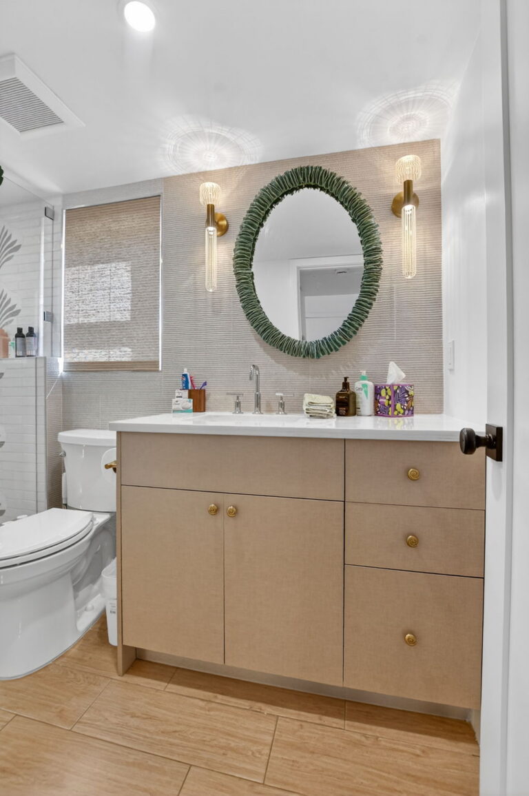 Alt text for Delray Cabana Condo: Bathroom — 1, 2 Photo 1: Bathroom vanity with wood-grain cabinet, white countertop, oval green-framed mirror, gold sconces, textured wallpaper, and woven wood blind.