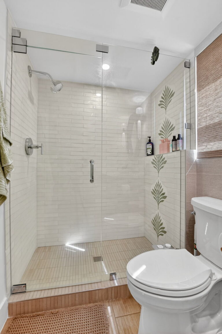 Walk-in shower with glass enclosure, white subway tile, palm-leaf accent strip, chrome fixtures, toilet, and wood-look floor tile.