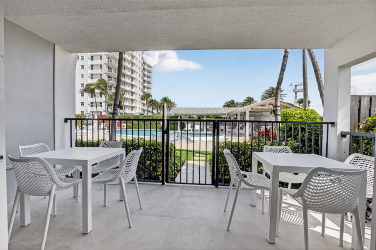 Private covered patio with grey tile flooring, two outdoor dining sets, and view of shared pool, palms, and condo buildings.