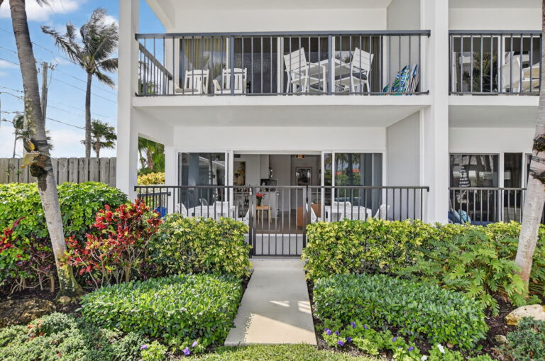 Ground-floor condo exterior with private covered patio, black metal gate, landscaped garden, upper balcony, and palm trees.