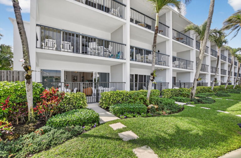 Three-story white condo building with black balcony railings, stepping-stone lawn path, tropical landscaping, and palm trees.