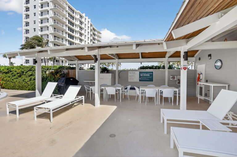 Covered community cabana with wood ceiling, white lounge chairs, outdoor dining tables, grill, and concrete floor.