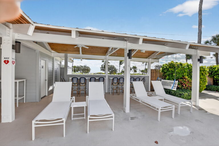 Covered patio with row of white lounge chairs, bar seating, porch swing, and lush green landscaping.