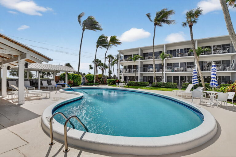 Shared community pool with kidney-shaped design, lounge chairs, striped umbrellas, and white condo building.