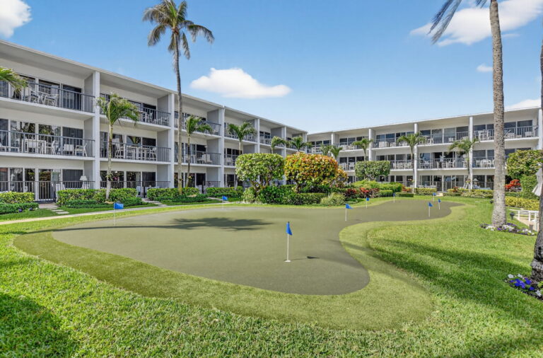 Wide view of courtyard putting green with synthetic turf, lawn, tropical plants, and surrounding white condo building.