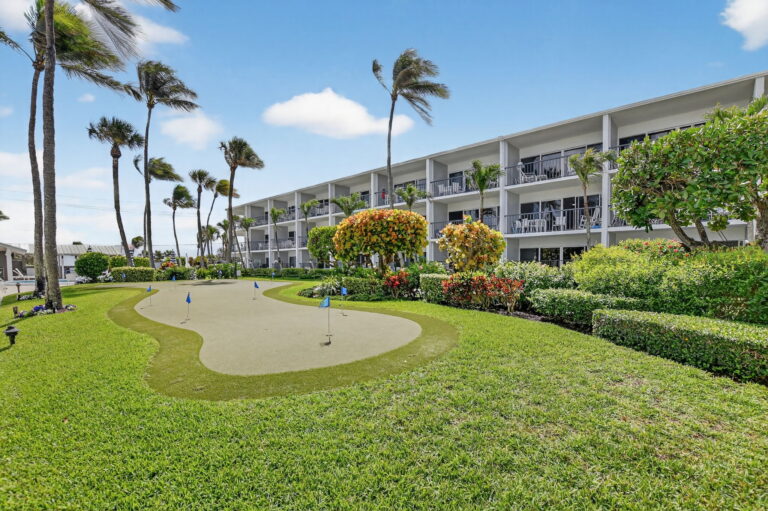 Close view of wavy putting green with orange flowering shrubs, palm trees, and white condo building in background.