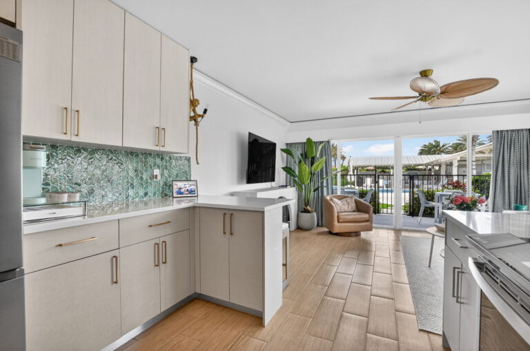 Kitchen view toward living area with leather armchair, wall-mounted TV, palm-leaf ceiling fan, and sliding doors to private patio.