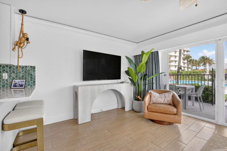 Living area with wall-mounted TV, white arched console, leather swivel chair, bird-of-paradise plant, and sliding doors to covered patio with pool view.
