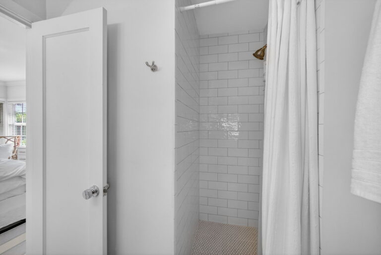 Open bathroom door showing white subway tile shower, gold showerhead, and white shower curtain.
