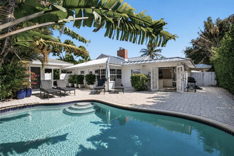 Wide backyard view with curved swimming pool, stone patio, grey lounge chairs, white home with French doors, and tropical palm landscaping