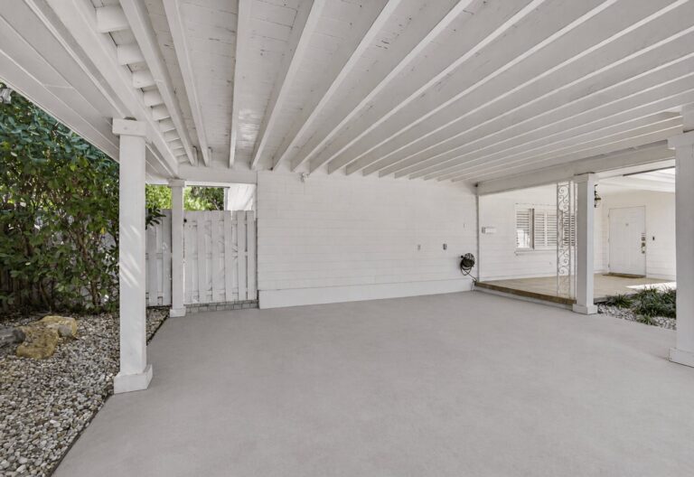 Covered carport with white beam ceiling, support columns, light grey concrete floor, and white picket gate at the rear.