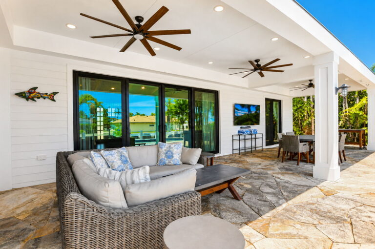 Covered patio with stone pavers, grey wicker sectional, ceiling fans, outdoor dining, fish wall art, and sliding doors.