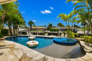 Pool deck view toward canal, circular spa, dock, and homes across the waterway under blue sky.