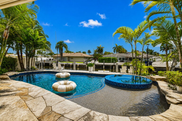 Pool deck view toward canal, circular spa, dock, and homes across the waterway under blue sky.