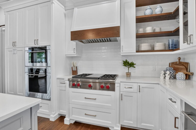 Kitchen cooking wall with stainless gas range, wood-trimmed vent hood, white subway tile backsplash, double wall oven, and open wood shelving with dishware.