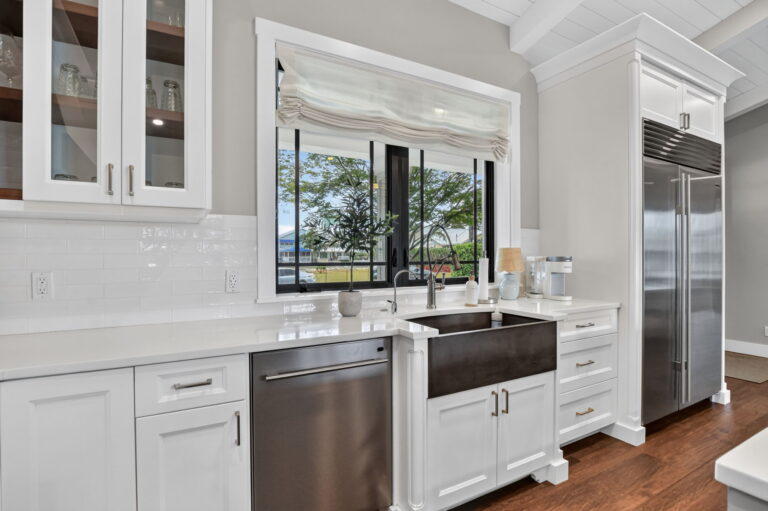 Kitchen sink area with farmhouse sink, black-framed window to greenery, built-in dishwasher, stainless refrigerator, and white cabinetry with glass-front uppers.