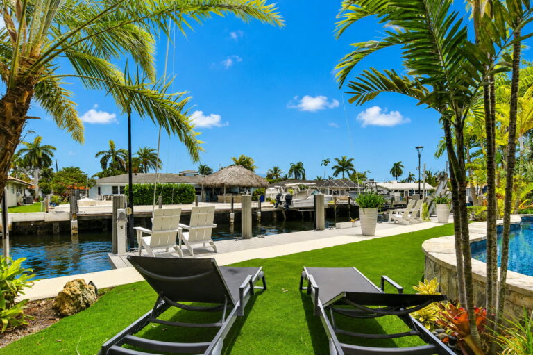 Canal-front lounge area with grey chairs on turf, private dock with boat lift, and palm trees.