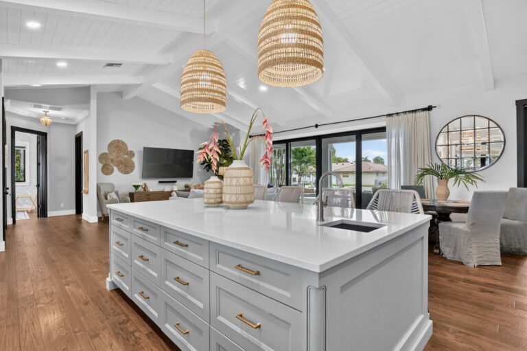 Kitchen island with white countertop, grey cabinets, gold hardware, woven pendant lights, vaulted ceiling, and open view to dining area and hallway.