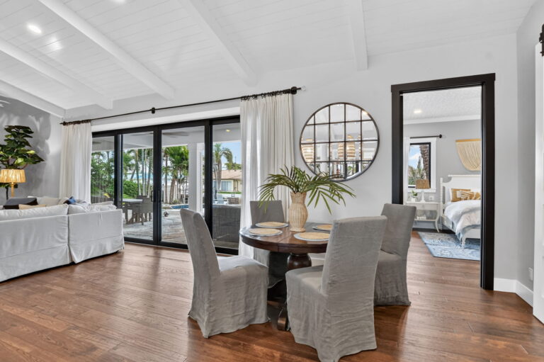Dining area with round wood table, grey slipcovered chairs, circular mirror, sliding doors to pool and patio, and open view to living room and bedroom doorway.